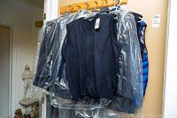 Photo shows multiple black and blue sleeveless vests hanging on a rack, some covered in plastic, located indoors near a wooden hook rack.