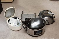Lot of four small kitchen appliances including Panasonic rice maker, KRUPS grill, Beaumark toaster, and Hamilton Beach cooker shown on beige carpeted floor.