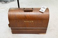 Front view of closed wood grain textured hard case with gold-tone SINGER logo and carry handle on top. Lot number tag attached.