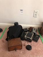 Group of cameras and accessories on floor including wooden box, camera bag, vintage camera, and flash unit.