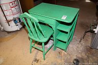 Photo showing green painted wood desk and matching chair from angled side view. Desk has drawer and two open shelves on right side. Both painted green with surface wear.
