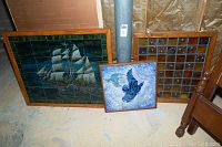 Three framed art pieces leaning against a wall on a floor; largest is a framed ceramic tile ship art, medium stained glass panel, and a smaller blue bird tile art in the middle
