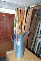Overall view of metal jug with wooden muddler sticks inside, placed on a small wooden surface with background items in storage.