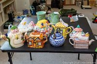 Overview shot of all nine ceramic teapots on black table showing a variety of whimsical designs and colors.