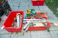 Red bins containing hand saws and smaller hand tools on pavement outdoors.