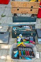 Wide shot showing a wooden workbench, green metal toolbox open with hand tools inside, and a plastic tray with assorted hand tools including screwdrivers.