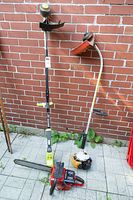 Overview of two string trimmers and a chainsaw leaning against brick wall