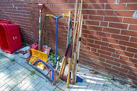 Overall view of assorted garden tools including shovels, hand saws, and a yellow snow shovel arranged against a brick wall on a paved surface.