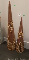 Photo showing two decorative cone-shaped Christmas trees made of brown woven material with gold bead embellishments, placed side-by-side against a wall on a wooden floor.