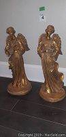 Pair of golden angel statues playing harps standing on floor against white wall.