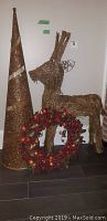 Full set showing tall glittery Christmas tree, wicker reindeer, and berry wreath on dark floor by white wall.