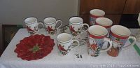 Photo of the full lot showing four small holiday coffee cups, four large mugs with Santa and Christmas patterns, and one red poinsettia-shaped plate on white cloth.
