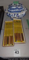 Photo of twelve fondue forks with wooden handles and colored tips in two boxes with original yellow packaging, placed next to blue and white ceramic soup tureen with lid.