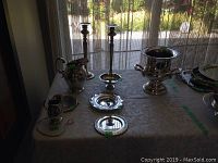 Silver items on display on a table with sheer curtains in background, including pitcher, candlesticks, trays, and a champagne bucket.