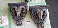 Pair of wall sconce shelves shown side by side on a cloth surface, displaying the ram's head and oak leaf detailed design with a bronze-like finish.