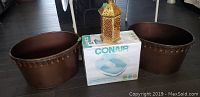 Two brown oval tin baskets, metal candle lantern, and boxed Conair foot spa on dark flooring under table.