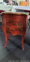Front view of red 2 drawer wooden end table showing Asian painted scenery on drawers and legs with wear and tear.