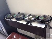 Four pieces of black cast cookware with glass lids on a shelf, including pots and skillets of varying sizes.