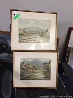 Photo showing two framed watercolor paintings by A. Widjaja placed vertically, both depicting rural mountain landscapes with fields and water.