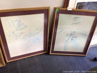 Two framed floral prints leaning against a wall on a dark floor