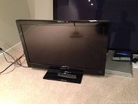 Full view of 32 inch Dynex television on a carpeted floor with remote on stand, screen off.