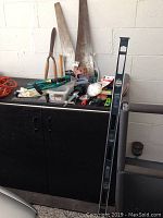 Photo showing assortment of carpentry tools on black cabinet including saws, level, hammer, small sledgehammer, hand tools, nails, and some hardware items.