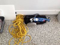Full view showing the electric polisher with black padded carrying case and long yellow rope, placed on a speckled floor.