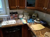 Kitchen counter full of various kitchen items including serving trays, glassware, Tupperware, folded table cloth, gloves, and a ceramic jar with fruit designs.