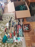 Collection of assorted garden tools laid out on a patterned carpet including loppers, pruners, snips, and gloves, with garden hoses in a box nearby.