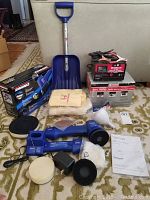 Full display of all items in the lot: cordless buffer, trolley jack, battery charger, emergency shovel, and cloths on carpeted floor.
