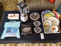 Full lot overview showing Oster blender, step stool, Pyrex dishes, decorative plates, and napkin rings boxed set on a coffee table.