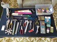 Wide view of assorted hand tools and hardware laid out on a black cloth, showing a variety of pliers, wrenches, hammers, scissors, and packaged hardware items.