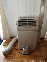 Front view of DeLonghi portable air conditioner with white body, air vent, exhaust hose, and window kit attached.