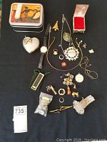 Photo showing full assortment of jewellery, watch, decorative boxes, lighter, and various pins and earrings laid out on black surface.
