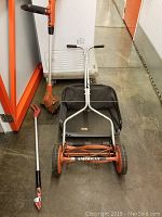 Full view of the orange American manual push mower with black grass catcher, orange and black Black and Decker line edger, and telescopic pruning shears on the floor.