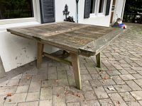 Weathered cedar outdoor dining table viewed from side, showing wood legs and table top with aged, weathered finish on a patio surface.