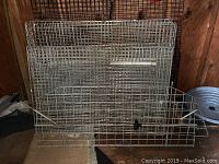 Three stacked metal wire animal cages leaning against a wooden wall on a carpeted floor, each about 3-4 feet in length, with hinged doors visible.
