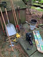 Wide view showing wooden handled garden tools including shovels, rake, hoes; yellow garden tool attachments on ground; stacked white and brown plastic plant pots; electric hedge trimmer and boxed root feeders.
