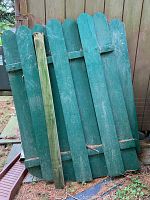Two weathered green wooden fence panels leaning against a wooden fence. Panels have rounded top slats and horizontal supports, one support is newer and unpainted.
