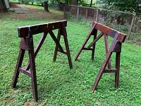 Pair of used wooden saw horses positioned on grass outdoors, showing their full frames and cross supports from a side angle