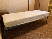 Full view of the blue metal cot with white mattress set on a carpeted floor.