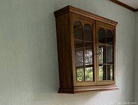 Front angle view of the wall mounted wood curio cabinet showing two glass decorated doors, crown molding and base molding.