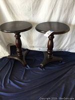 Matching pair of dark wood end tables with round tops, turned central pedestal, and ball feet on triangular bases, shown on blue cloth against white backdrop.