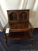 Front view of wooden end table with mid-century style, lyre wire doors, glass inset top, turned legs with casters.