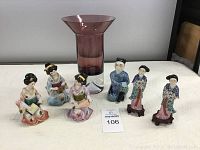 Photo showing six ceramic Asian figurines arranged in front of a tall purple/pink glass vase, figurines are approx. 6 inches tall