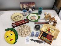 Overview of assorted travel souvenirs including plates, mugs, wooden items, novelty decor, and kitchen accessories arranged on a white fabric surface.