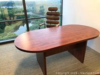 Large oval meeting table with wood panel ends shown near window with outdoor scenery. The surface has a clean medium brown wood finish with a slight shine. The table rests on two solid panel legs forming an End panel style.