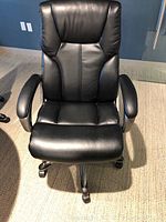 Front view of black leather-like padded office chair with high backrest, padded armrests, and caster wheels on a carpeted floor.