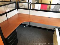 L-shaped cubicle desk with attached cubicle partition walls. Wood finish desk surface with cable grommets, black metal frame, and filing cabinet beneath.