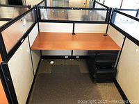 Cubicle desk with attached cubicle partitions and black under-desk pedestal drawers, on carpet flooring.